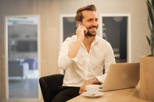 a man in a white button-up shirt smiling whilst on the phone in front of a laptop